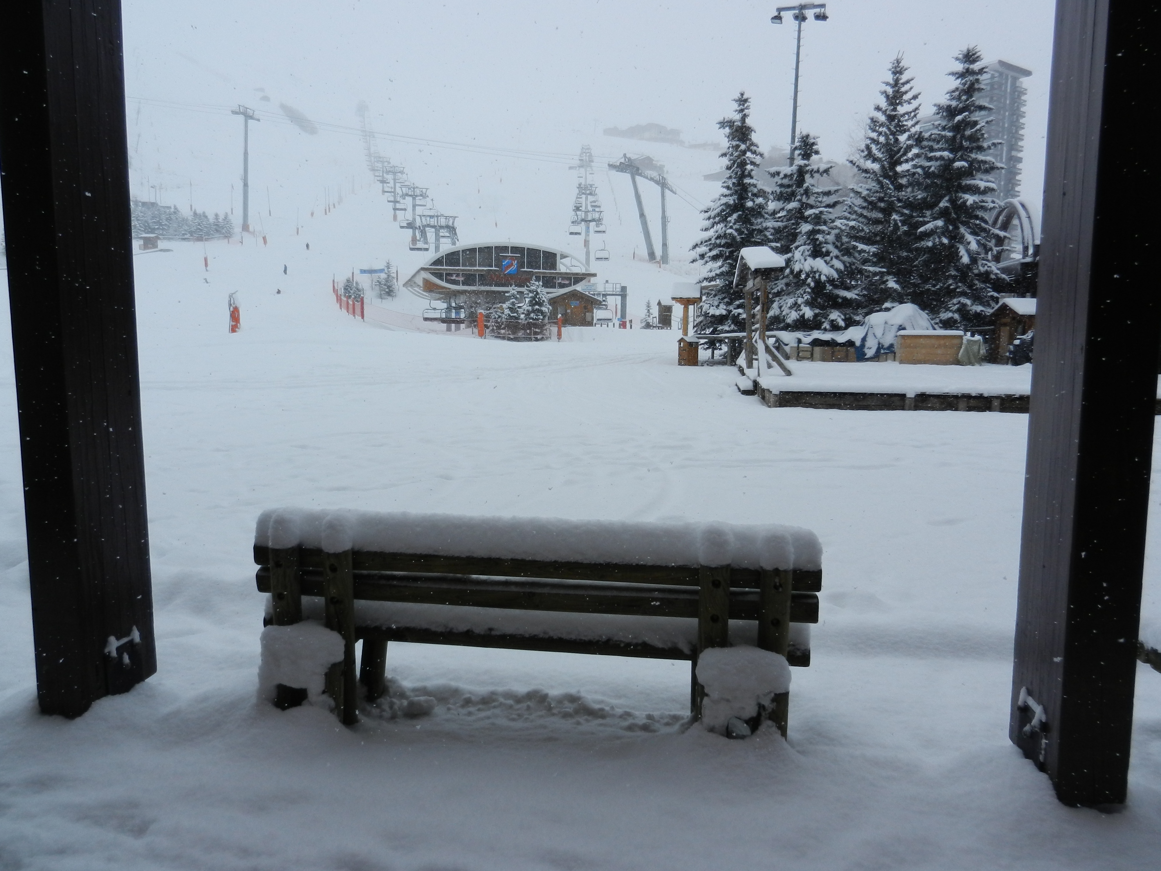 Wooden bench covered with a thick layer of snow facing the slopes and ski lifts of a ski resort during snowfall.